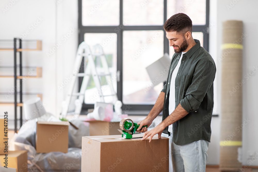 moving, people and real estate concept - happy smiling man with adhesive tape packing box at new home moving, people and real estate concept - happy smiling man with adhesive tape packing box at new home