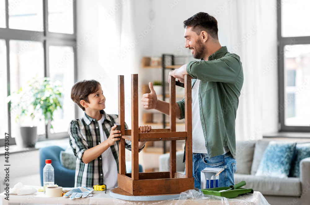 furniture renovation, diy and home improvement concept - happy smiling father showing thumbs up to his son sanding old round wooden table with sponge at home