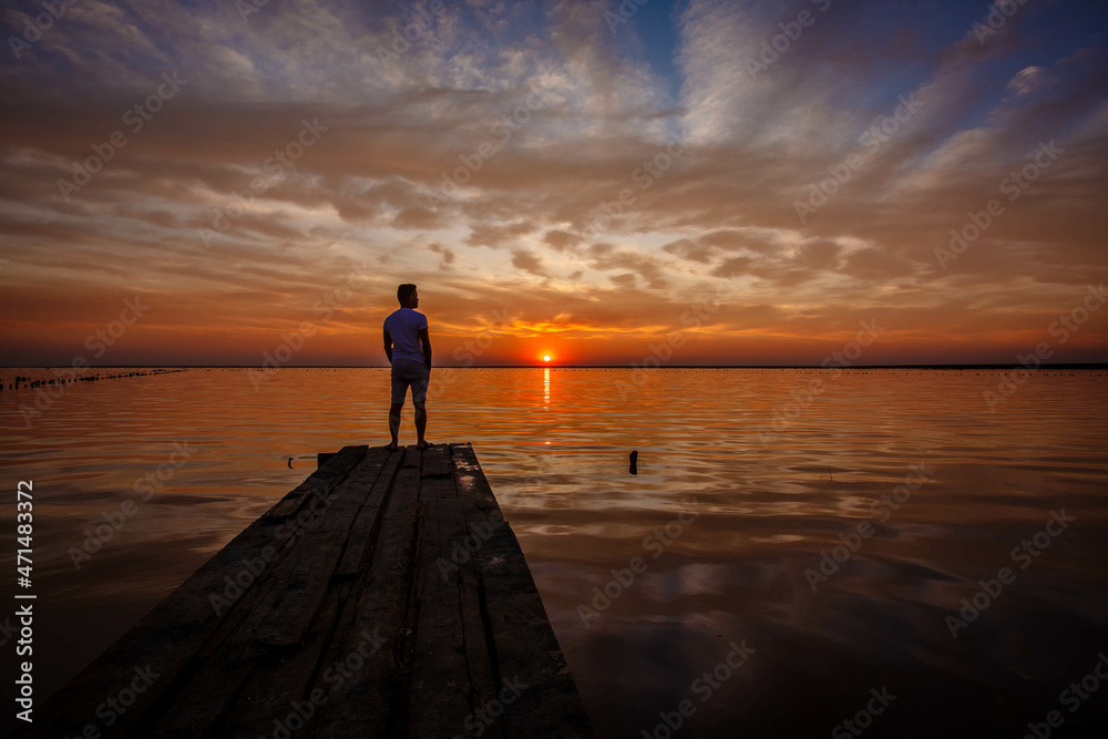 Fototapeta premium A man stands on a wooden pier and looks at the horizon of the sunset of the lake.