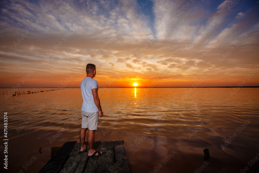 A man stands on a wooden pier and looks at the horizon of the sunset of the lake.