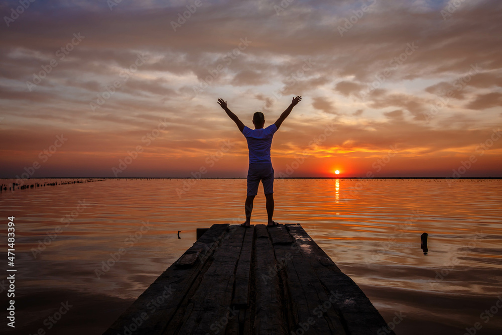 A man with raised arms stands and looks at the sunset on the lake.