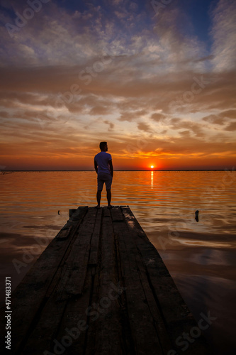 A man stands on a wooden pier and looks at the horizon of the sunset of the lake.