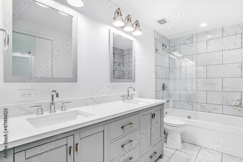 An elegant, remodeled bathroom with a grey vanity and bronze hardware. The shower has a large shower head and marble tiles and glass wall line the sides.