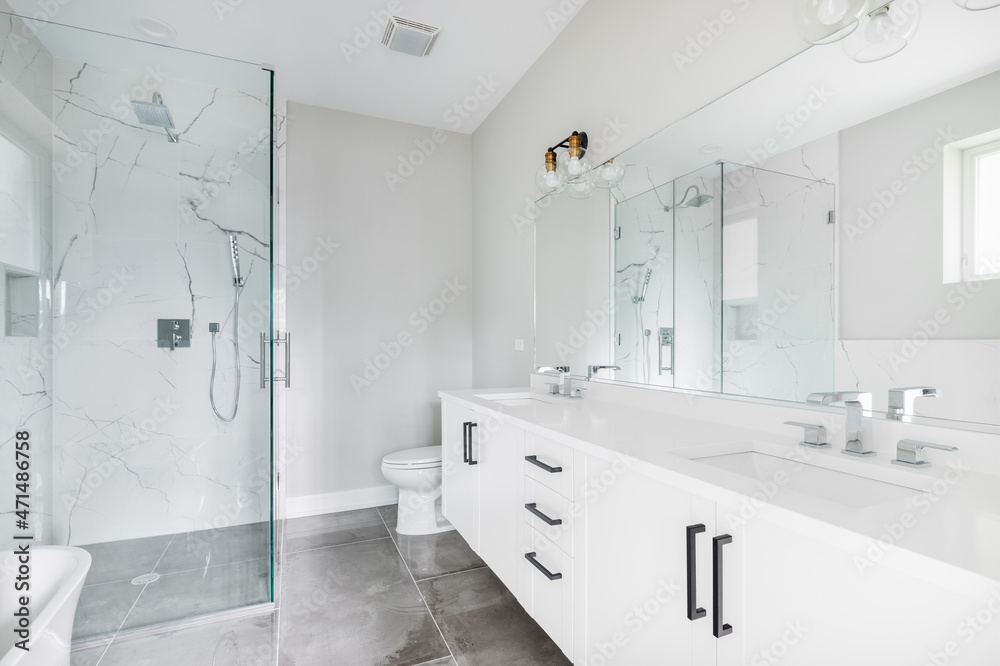 A modern, white luxury bathroom with black hardware and chrome faucets. A  large glass walled shower sits in the corner with marble tiles from floor  to ceiling. Stock Photo | Adobe Stock