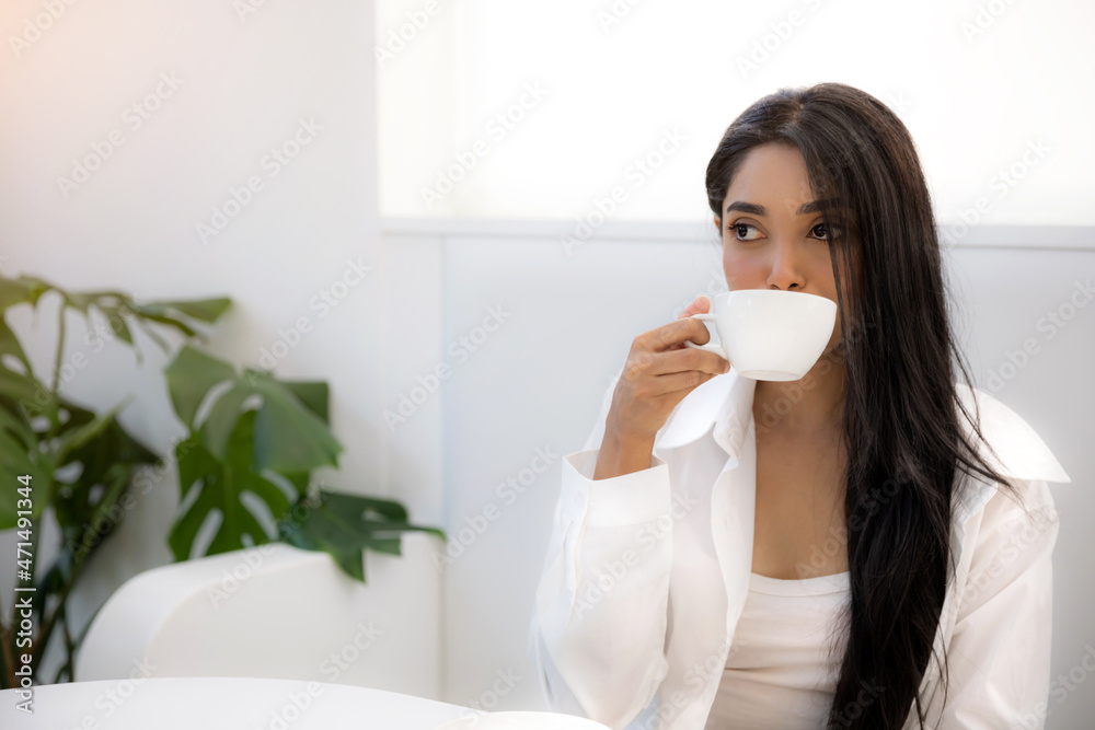 Portrait of young Indian woman drinking coffee or tea at table in cafe or office or stylish home or medical centre waiting Portrait of young Indian woman drinking coffee or tea at table in cafe or office or stylish home or medical centre waiting