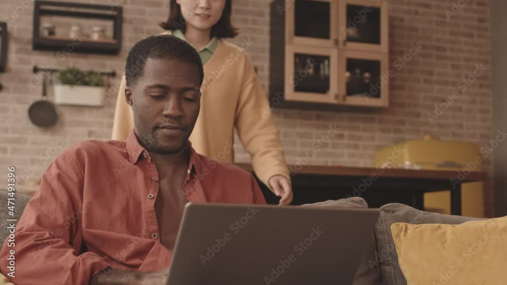 Waist up slowmo shot of young African-American man surfing the Internet on laptop sitting on sofa in modern apartment sharing news with his Asian girlfriend Waist up slowmo shot of young African-American man surfing the Internet on laptop sitting on sofa in modern apartment sharing news with his Asian girlfriend