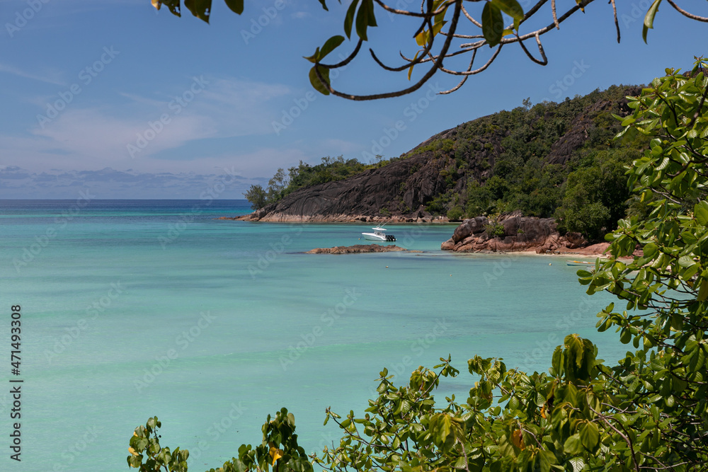 A view from high point to granite coast of a tropical island. Shore covered with jungle. A yacht is moored in the blue lagoon. In the foreground is the blurry green foliage of tropical trees.