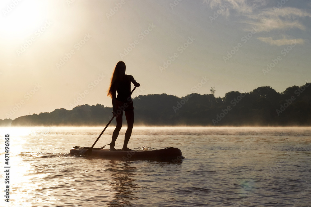 Silhouette of young woman paddle boarding at dawn in the morning haze in the river