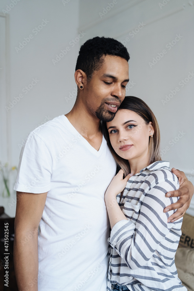 Multiracial couple in love embraces in cozy living room at home. A close-up portrait of smiling caucasian woman and african man are hugging. Love and affection concept Multiracial couple in love embraces in cozy living room at home. A close-up portrait of smiling caucasian woman and african man are hugging. Love and affection concept