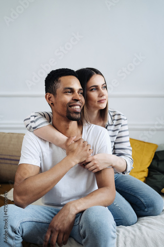 Portrait of happy young African Caucasian couple hug sitting on couch at home, smiling mixed race husband and wife spend time together, embrace relaxing on sofa. Healthy relationships concept