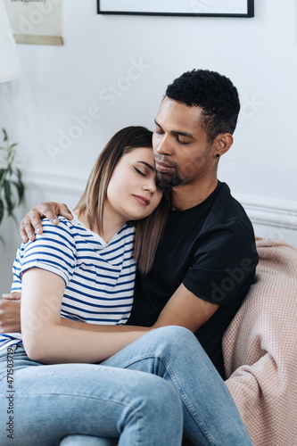 Portrait of happy young African Caucasian couple hug sitting on armchair at home, smiling mixed race husband and wife spend time together, embrace relaxing. Healthy relationships concept