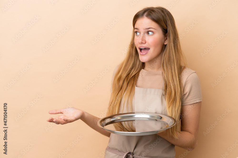 Young blonde waitress woman with tray isolated on beige background with surprise expression while looking side Young blonde waitress woman with tray isolated on beige background with surprise expression while looking side