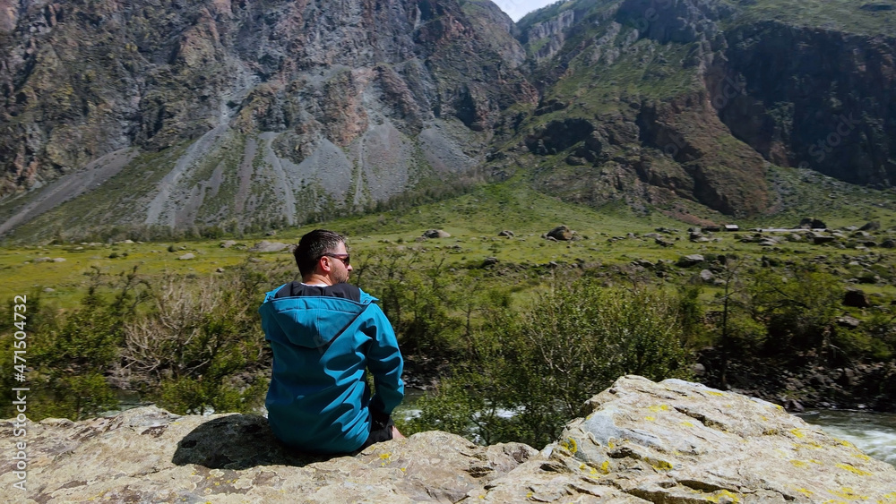 A man with glasses sits on the edge of a cliff wearing a blue anorak. Katu-Yaryk canyon Chulyshman Valley. Altai