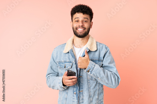 Young Moroccan man using mobile phone isolated on pink background giving a thumbs up gesture © luismolinero