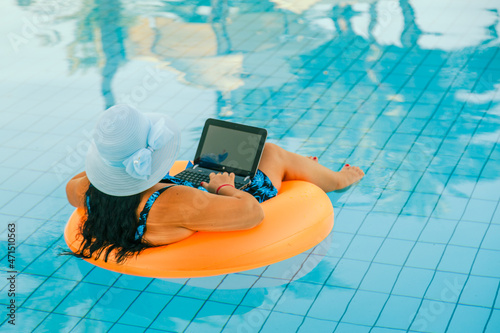 Brunette woman in a sun hat in the pool in a swimming circle with a cocktail and a laptop with her back to the camera.