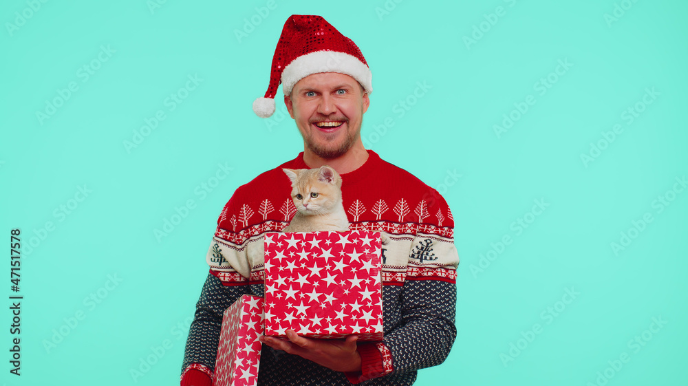 Man in Christmas red sweater hat smiling, unwrapping gift, opening box with pet cat and expressing great surprise. Young fashion guy indoors studio shot isolated on blue background. Happy New Year