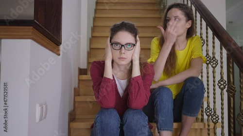 Tired woman in eyeglasses covering ears with hands sighing as excited twin sister talking sitting on stairs at background. Portrait of skeptic intelligent Caucasian nerd with cheerful sibling at home