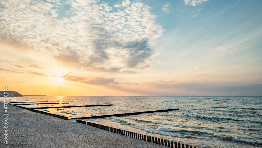 Panorama of the evening sea with a wooden breakwater by the sandy shore against the background of the sunset
