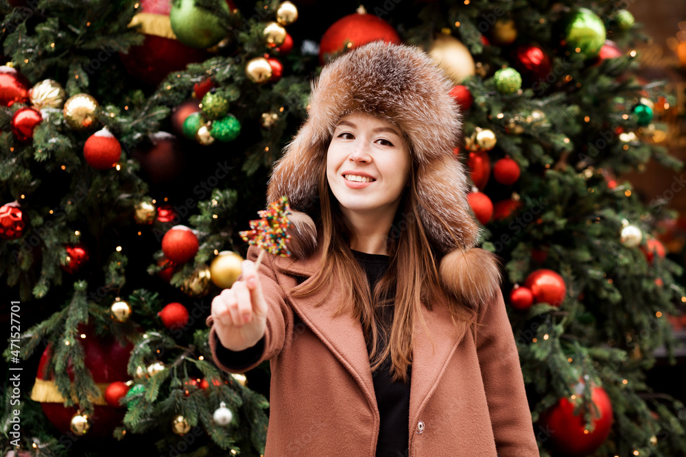 Attractive woman in hat with ear flaps is showing lollipop standing in front of Christmas tree. 