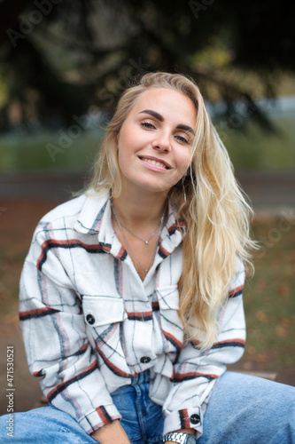 pretty caucasian young blonde woman in white flannel shirt is sitting on the branch in the autumn park