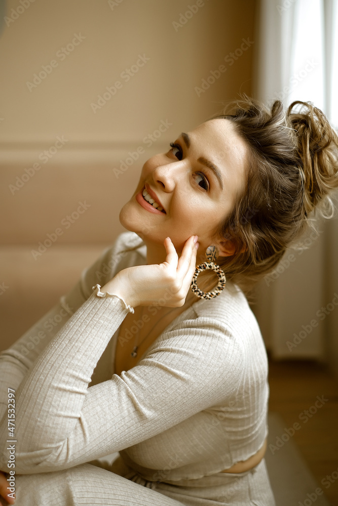 portrait of young caucasian woman in a beige house suit sitting on the floor, a beautiful lady with large earrings and high hairstyle in a stylish apartment, a stylish image of a young successful portrait of young caucasian woman in a beige house suit sitting on the floor, a beautiful lady with large earrings and high hairstyle in a stylish apartment, a stylish image of a young successful