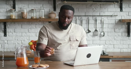 African male freelancer eats cookies and works at the computer while sitting at the kitchen table at home.
