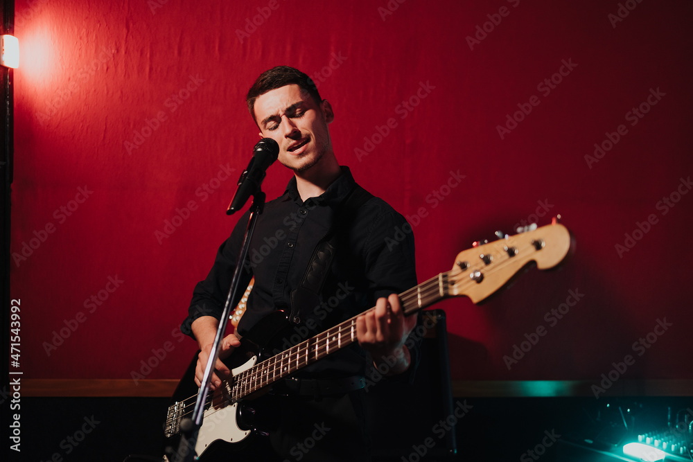 Obraz premium Talented handsome young guitarist man singing a song in studio recording on red background surrounded by instruments. Passion, hobby, singer, electric guitar