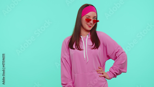 Portrait of seductive cheerful teen girl in pink hoodie wearing sunglasses, charming smile, blinking eye. People emotions. Young lovely adult woman posing isolated on blue studio wall background