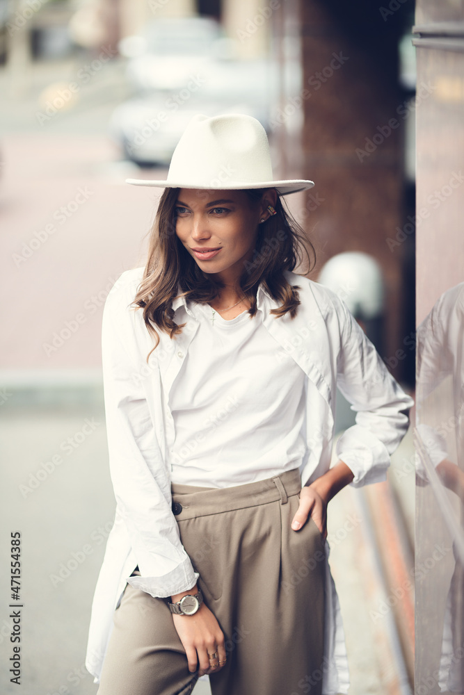 Adorable beautiful young woman expresses happiness, wears fashionable clothes and hat, poses on street, against buildings