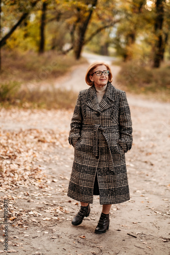 Beautiful and slim elderly woman in a woolen coat, scarf posing, standing under a beautiful tree with golden leaves in an autumn park, relaxing and enjoying the bright foliage, happy retirement