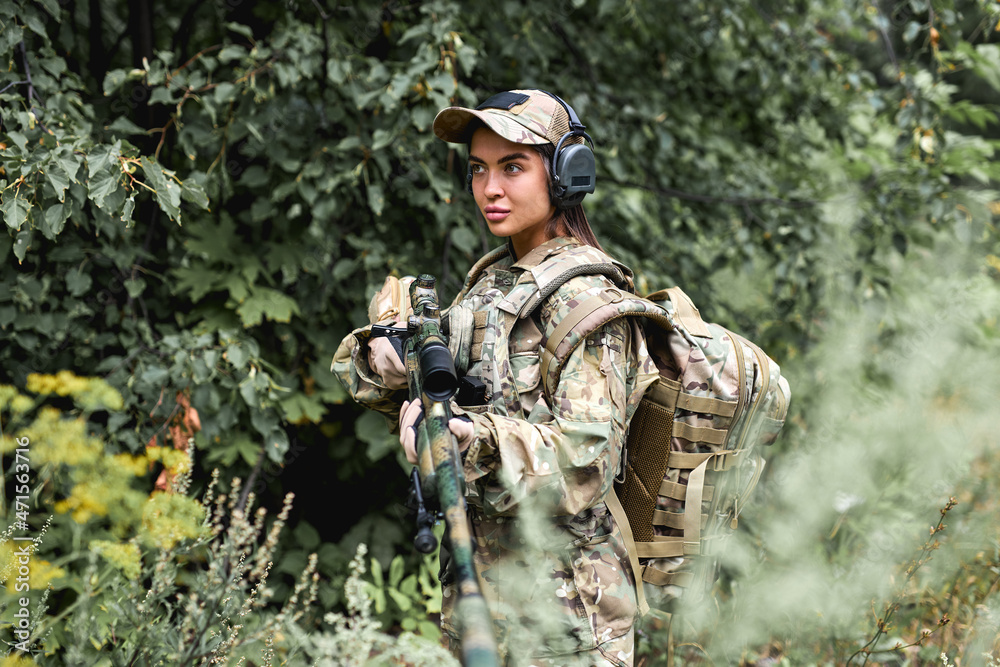 Caucasian Military lady woman in tactical gear posing for photo in forest during summer. Wearing green camo uniform and assault rifle, in military gear and headset, lady is looking at side