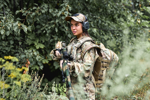 Caucasian Military lady woman in tactical gear posing for photo in forest during summer. Wearing green camo uniform and assault rifle  in military gear and headset  lady is looking at side