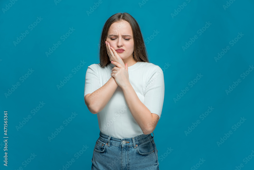 Young sad unhappy girl suffer toothache, pain from sensitive tooth on blue background. Dental problems, caries