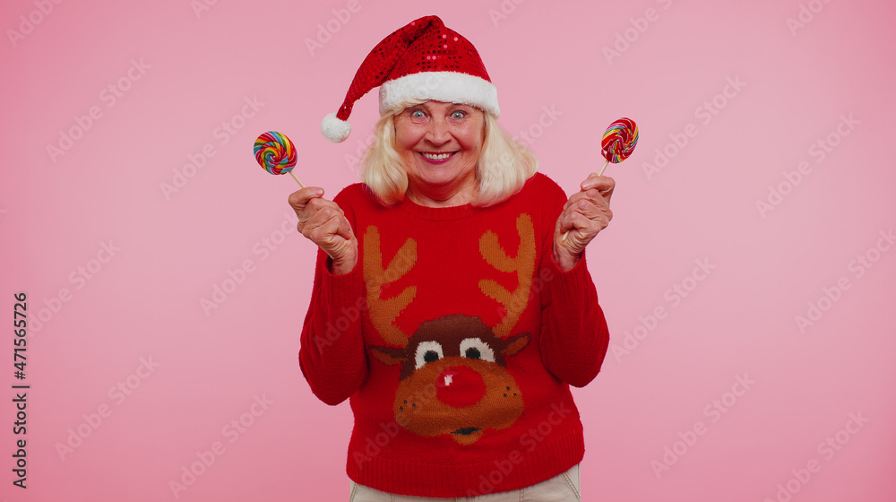 Grandmother woman in New Year sweater, hat holding candy striped lollipops, hiding behind them fooling around dancing isolated on pink studio wall background. Happy Christmas celebration merry holiday