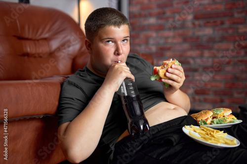 Overweight boy with fast food on floor near sofa at home  eating junk food drinking sweet beverages  lazy fat teenage boy in casual wear at home alone  having no control in eating behavior.