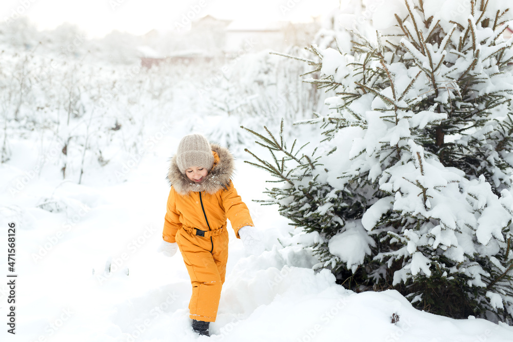 A child in a yellow jumpsuit walks among fir trees in snowdrifts on a frosty cold day. The girl rejoices at the first December snowfall. Winter children's games and entertainment