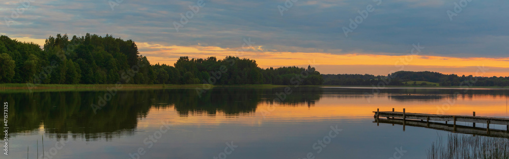 Panoramic view of the lake at sunset. A wooden footbridge enters the lake. There is a reflection of trees and clouds in the water