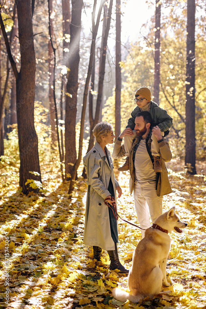 Fototapeta premium Da, mom and child boy walking with dog in warm sunny day in forest during leaf fall at autumn season. Lovely and carefree caucasian family consisted of parents, child and pet enjoy the walk