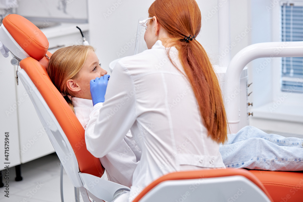 Child girl patient examining by female dentist with tools in dental clinic. Rear view on professional redhead woman dentist in white uniform holding checkup for caucasian child, treatment Child girl patient examining by female dentist with tools in dental clinic. Rear view on professional redhead woman dentist in white uniform holding checkup for caucasian child, treatment