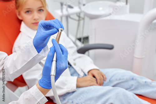 Healthy teeth and smile. Female doctor sitting at chair, holding dental drill in hand. Side view on young woman in white medical uniform going to treat teeth of child girl, close-up hands with tool