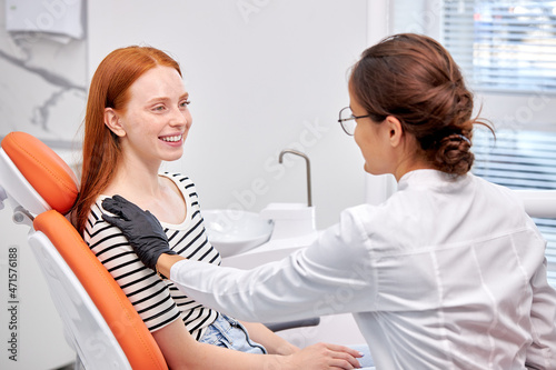 Side View On Young Professional Female Dentist Calming Patient Down Before Treatment, Attractive Redhead Caucasian Woman Is Smiling, Having Nice Talk, Discussing. Healthcare, medicine concept
