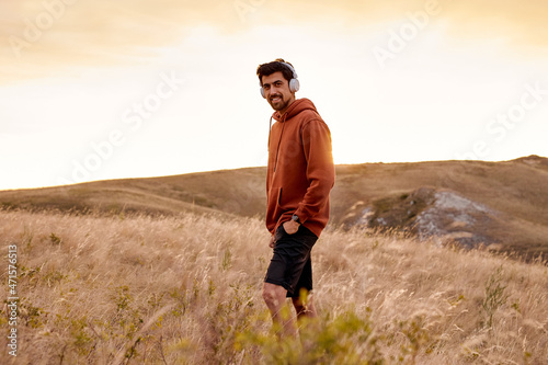 male walking alone in field listening to music, at summer or autumn season, handsome brunette guy in red casual hoodie having rest in countryside, healthy and fit male thinking, in contemplation