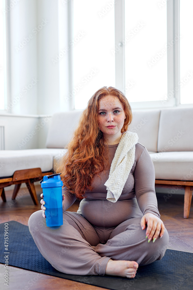 plus size woman with red hair siit on fitness mat drinking water, after training workout at home to lose weight and get strong flexible body, exercising on mat, looking at camera, posing plus size woman with red hair siit on fitness mat drinking water, after training workout at home to lose weight and get strong flexible body, exercising on mat, looking at camera, posing