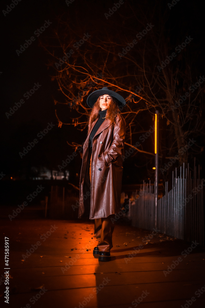 Girl in a hat in a portrait at night in a raincoat on a parcel. Caucasian brunette model looking at camera