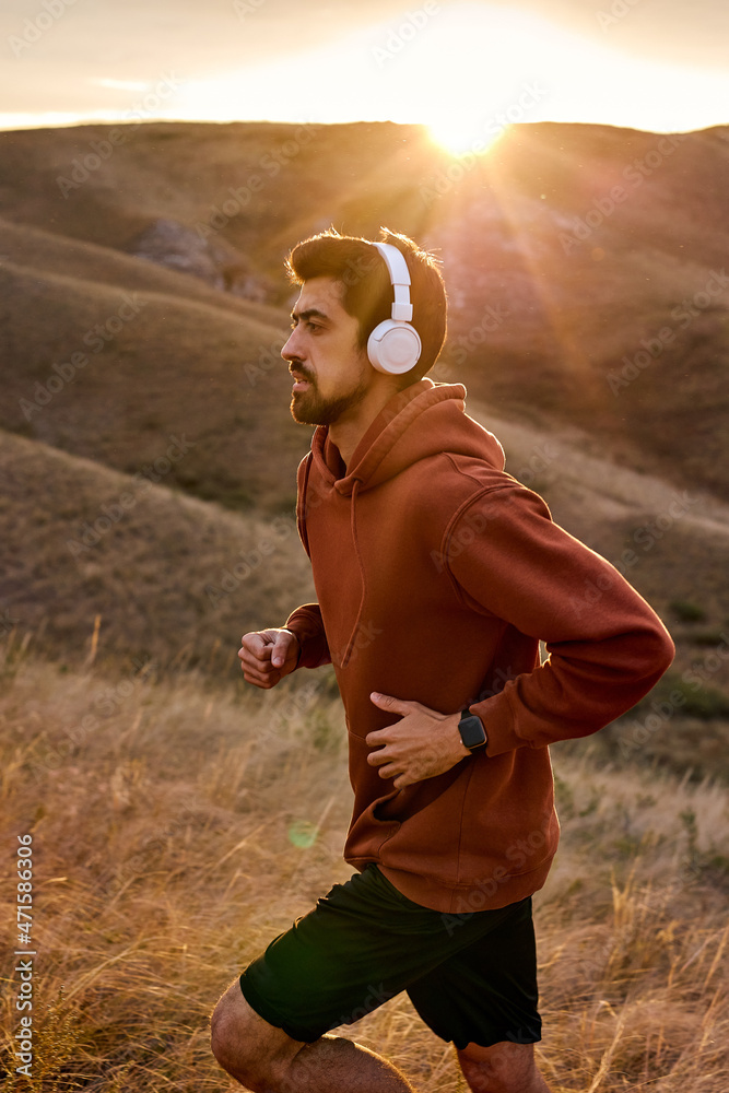 Man jogging on field. Male runner running listening to music on smartphone wearing headphones. Young caucasian guy fitness sport model in red casual sportive hoodie. Sport and healthy lifestyle