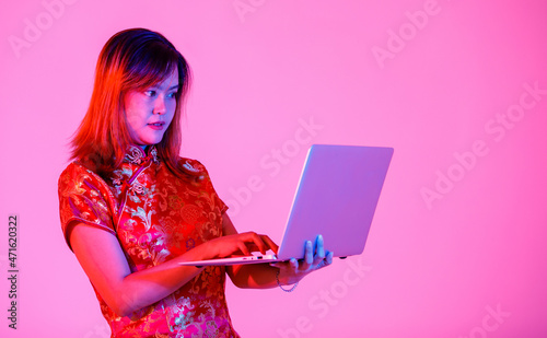 Studio shot of millennial Asian young teen female model in beautiful red Chinese traditional cheongsam qipao shirt sitting holding laptop computer surfing shopping online on pink light background photo