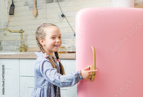 A five-year-old girl in a pink coat opens an old vintage pink refrigerator in a retro-styled kitchen.