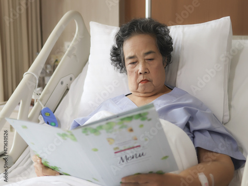 Asian senior female patient  sitting and reading food menu in hospital bed, ordering food. Elderly health concept.