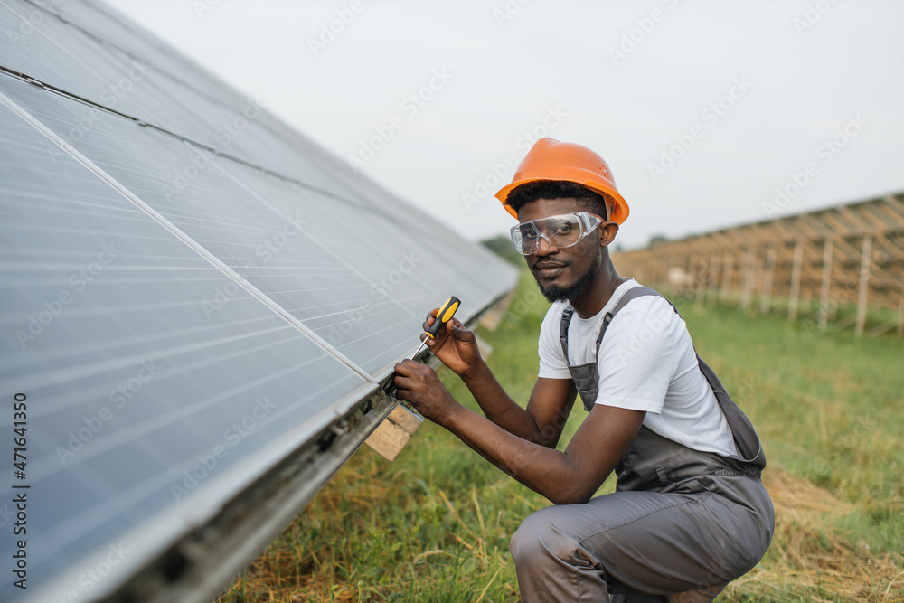 African american man in safety helmet and glasses tighten nuts on solar panels with screwdriver. Competent technician using tools while performing service work on station.