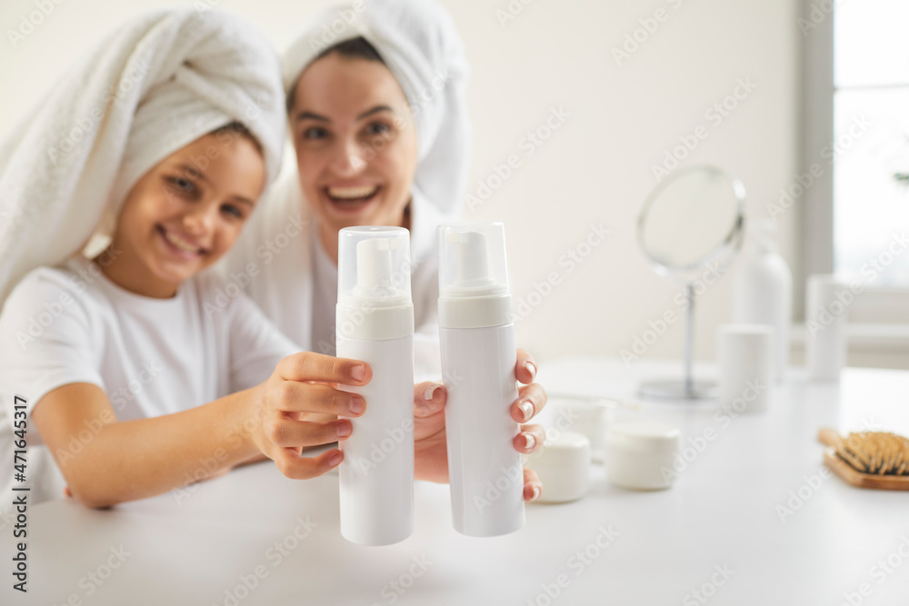 Beautiful family doing morning hair care. Smiling mom and kid in bathroom towels show clean plastic mockup shampoo, conditioner or face wash bottles. Children and adults' dermatology products concept Beautiful family doing morning hair care. Smiling mom and kid in bathroom towels show clean plastic mockup shampoo, conditioner or face wash bottles. Children and adults' dermatology products concept